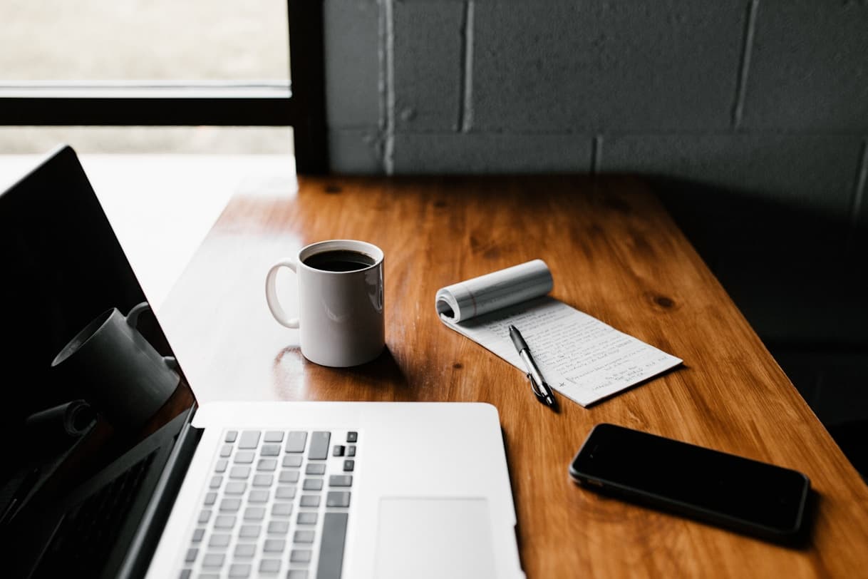 Image of a work desk with a laptop, pen, phone, coffee, and notepad on it